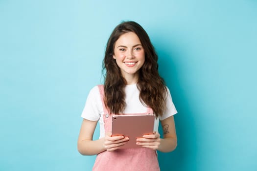 Young smiling female model holding pink digital tablet in hands, looking friendly at camera, assisting you, standing over blue background.