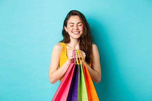 Young beautiful woman daydreaming about wearing new clothes, holding shopping bags, close eyes and smile dreamy, standing over blue background.