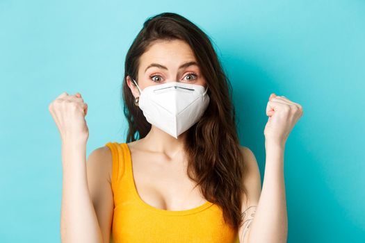 Health, covid-19 and lockdown concept. Excited young woman in respirator mask, saying yes, express rejoice and triumph, making fist pump from good news, standing against blue background.