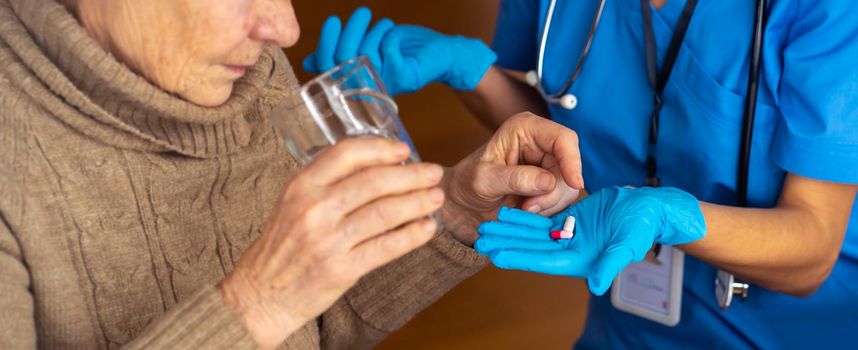 A young girl nurse with a stethoscope holds medication for an elderly patient, age 80, in her hand. A retired woman is holding a glass of water and is going to drink vitamins to improve health.