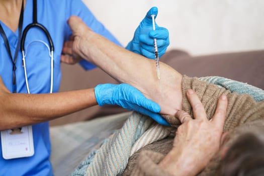 The nurse makes an injection with the vaccine to the patient at home. A young doctor in a blue suit holds the hand of an elderly woman and makes injection with a syringe. Medicine and healthcare.