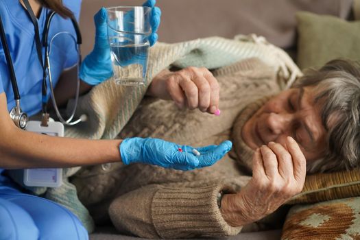 A young nurse is caring for an elderly 80-year-old woman at home. She holds a glass of water and gives medicine pills a pensioner retired woman who lies and rests in bed.