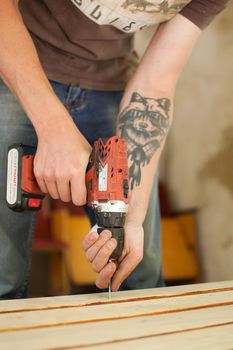 Ukraine, Goshcha, voluntary event, - August, 2021 - skilled young male worker is using power screwdriver drilling during construction wooden bench, do it yourself.