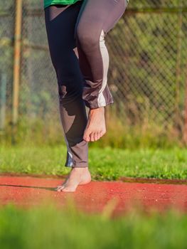 Middle age woman stretching knees and ankles, she jumping bare feet on red rubbery running track of outdoor stadium.