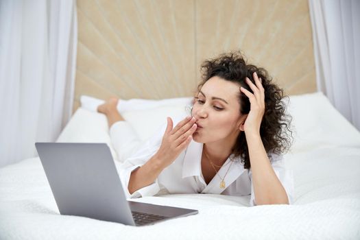 Curly woman using laptop computer sitting on white bed browsing or chatting with friends online