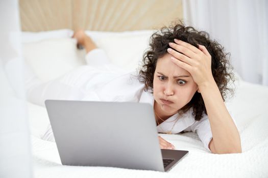 Curly woman using laptop computer sitting on white bed browsing or chatting with friends online