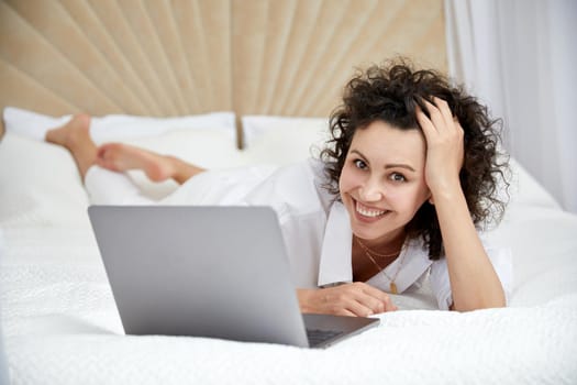 Curly woman using laptop computer sitting on white bed browsing or chatting with friends online