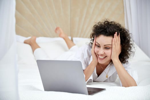 Curly woman using laptop computer sitting on white bed browsing or chatting with friends online