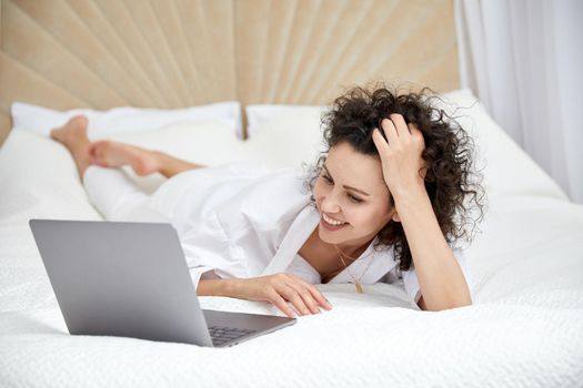 Curly woman using laptop computer sitting on white bed browsing or chatting with friends online