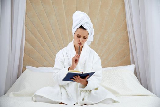 woman sits on bed wearing bathrobe after shower, makes notes in diary. Blogger creats publication for blog in notebook