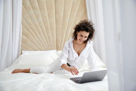 Curly woman using laptop computer sitting on white bed browsing or chatting with friends online