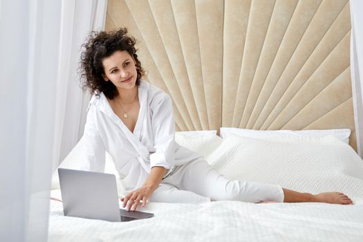 Curly woman using laptop computer sitting on white bed browsing or chatting with friends online