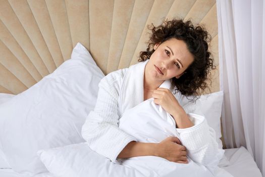 Sad depressed curly woman hugging pillow sitting in white bed alone