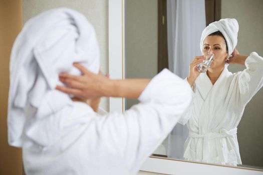 Beautiful young woman drinking pure water standing in front of mirror in the morning. Concept of healthy lifestyle