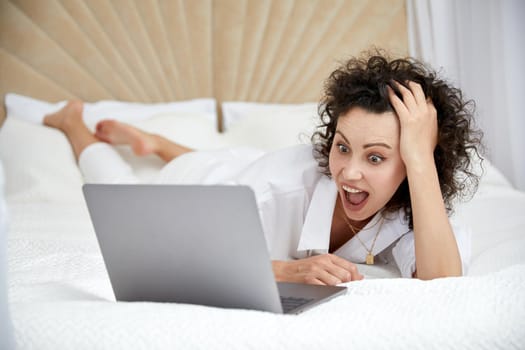 Curly woman using laptop computer sitting on white bed browsing or chatting with friends online