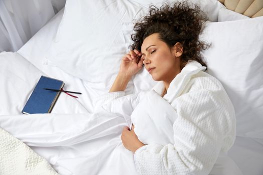 Curly woman sleeping in bed with notebook near by wearing white bathrobe