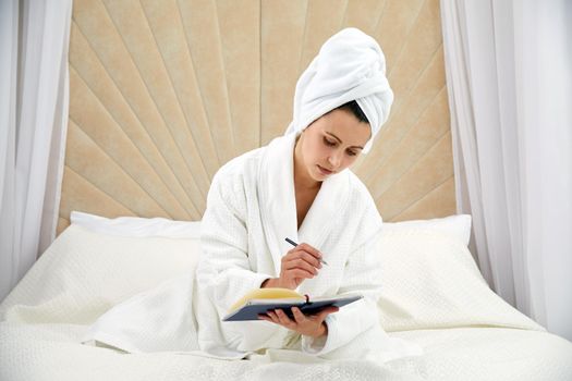 woman sits on bed wearing bathrobe after shower, makes notes in diary. Blogger creats publication for blog in notebook