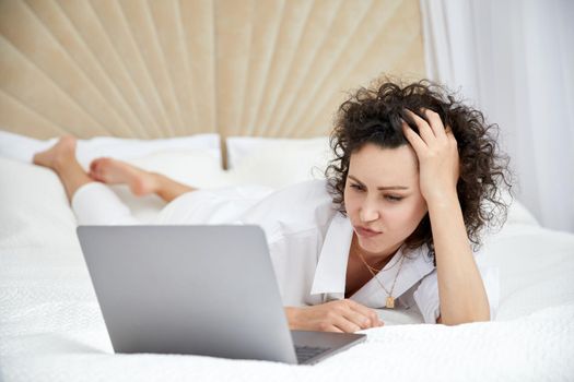 Curly woman using laptop computer sitting on white bed browsing or chatting with friends online