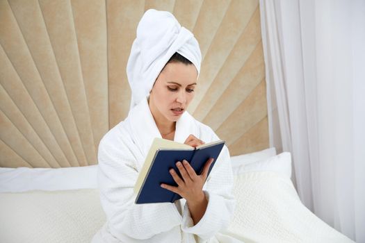 woman sits on bed wearing bathrobe after shower, makes notes in diary. Blogger creats publication for blog in notebook