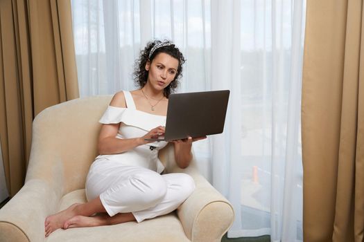 Beautiful young woman using laptop sitting in armchair. Concept of relaxed business lady