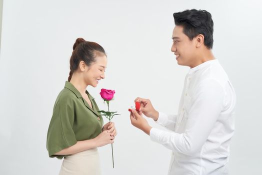 Attractive young woman is standing with roses in hands while handsome man is making proposal with ring in hands.