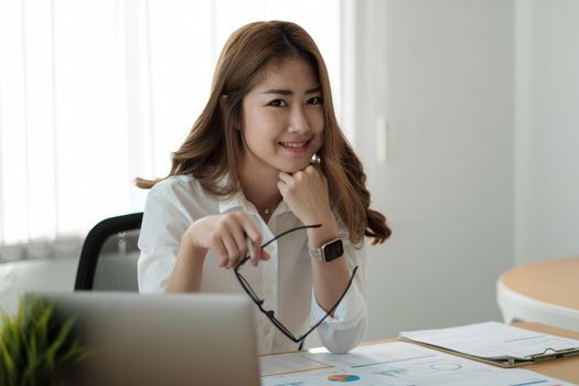 Portrait young beautiful smiling asian business woman holding a glasses and laptop Placed at the wooden table at the office,