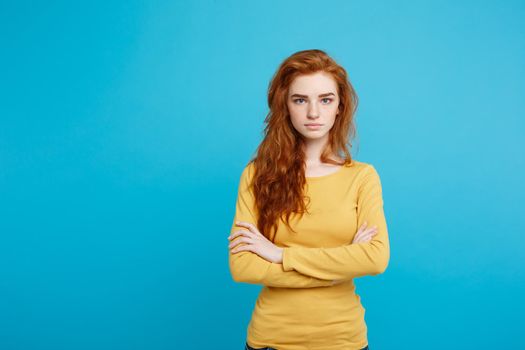 Portrait of young beautiful ginger woman with tender serious face crossing arms looking at camera. Isolated on pastel blue background. Copy space.