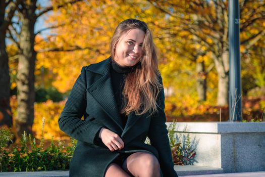 A woman with long hair sits on a bench in an autumn park and laughs. Autumn season.