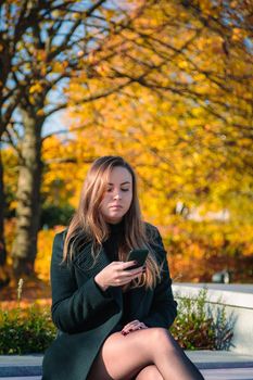 A woman with long hair sits on a bench in an autumn park and looks into a mobile phone. Autumn season.