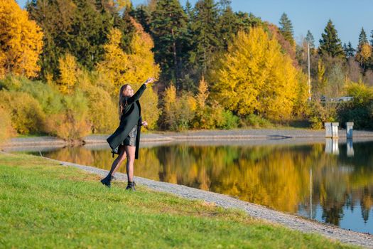 A woman with long hair walks in an autumn park by a pond and throws stones into the water. Autumn season.