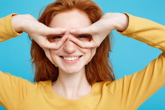 Fun and People Concept - Headshot Portrait of happy ginger red hair girl with freckles smiling and making finger glasses. Pastel blue background. Copy Space.