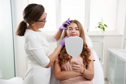 Cosmetologist using facial marker on patient face before beauty treatment in spa salon. Client is looking into mirror in in beauty clinic