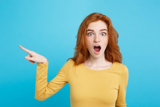 Fun and People Concept - Headshot Portrait of happy ginger red hair girl with pointing finger away and shocking expression. Pastel blue background. Copy Space.