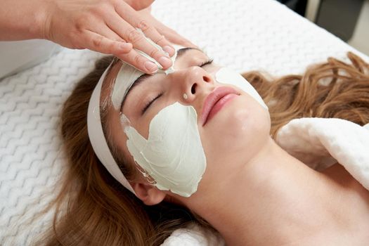 Cosmetologist applying rejuvenating clay facial mask on a female face in beauty salon. Facial treatment concept