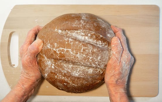 Black fresh round rye bread in the hands of an elderly man on the kitchen board.  Bread in flour