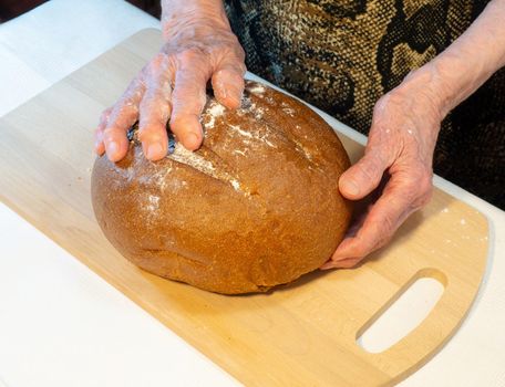 Black fresh round rye bread in the hands of an elderly woman
 on the kitchen board. Bread in flour