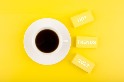 Hot trends 2022 written on rectangles next to cup of coffee on bright yellow background. Concept of newest, latest, hot and popular trends of upcoming 2022 year