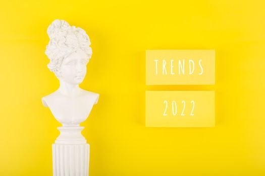 Minimal flat lay with words Trends 2022 written on two rectangles on bright yellow background next to woman gypsum statue. Concept of newest, latest, hot and popular trends
