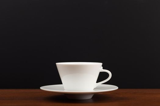 Selective focus, close up of white ceramic coffee or tea cup with saucer on brown wooden table against dark black background with copy space.