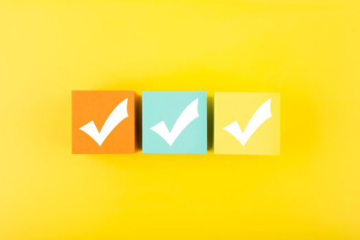 Three checkmarks on multicolored toy cubes on bright yellow background. Concept of questionary, kids related checklist, to do list, planning, business or verification.