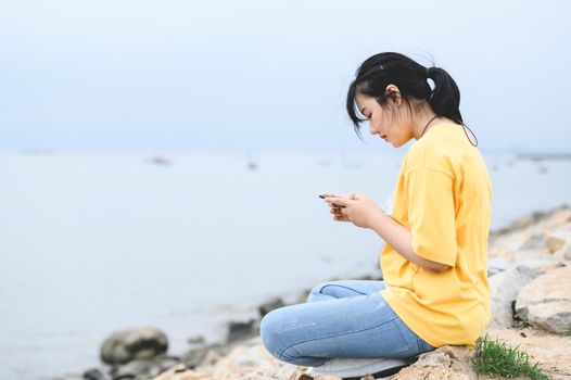Young Asian woman is using a smartphone at the seaside with her face mask removed to breathe fresh air after vaccination on Covid-19 or Coronavirus epidemic. People lifestyle health concept.