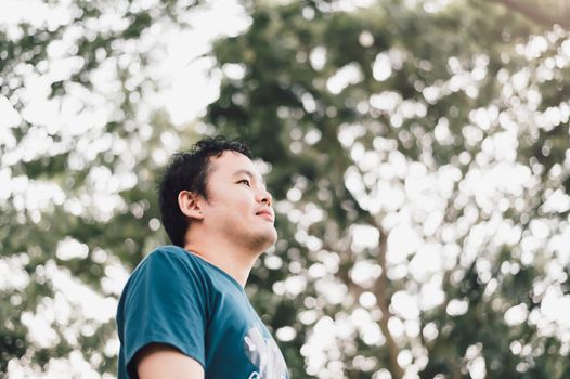 Asian man standing smiling happily under big tree in the public park. Nature and people lifestyle. Healthy single man having success life concept. Copy space