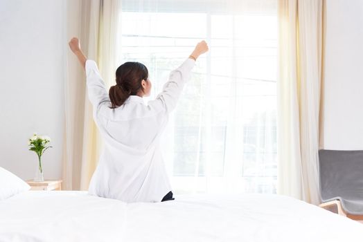 Back view of woman stretching in morning after waking up on bed near window. Holiday and Relax concept. Lazy day and Working day concept. Office woman and worker in daily life theme