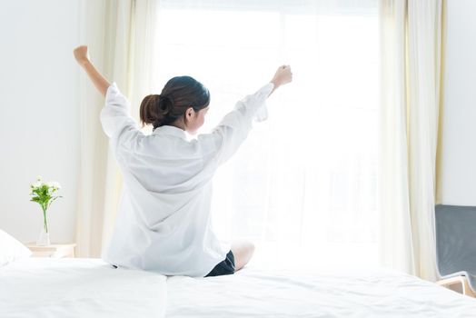 Back view of woman stretching in morning after waking up on bed near window. Holiday and Relax concept. Lazy day and Working day concept. Office woman and worker in daily life theme