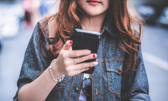 Close up of Beauty woman in jeans jacket using smart phone while walking on street. People and Lifestyles concept. Technology and communication theme. Selective focus on shirt. Blurry background.