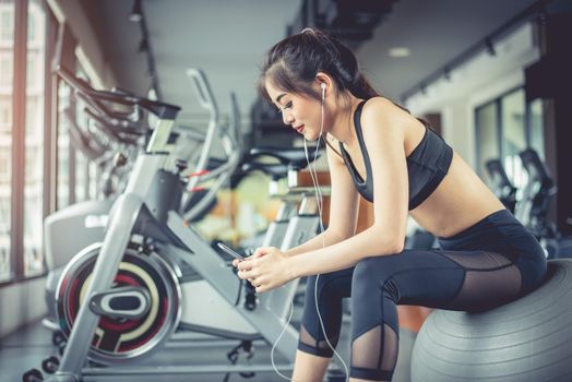 Asian woman sitting on fitness ball and listening music in fitness training gym with smart phone. Relax and sport workout training concept. Technology and Entertainment theme