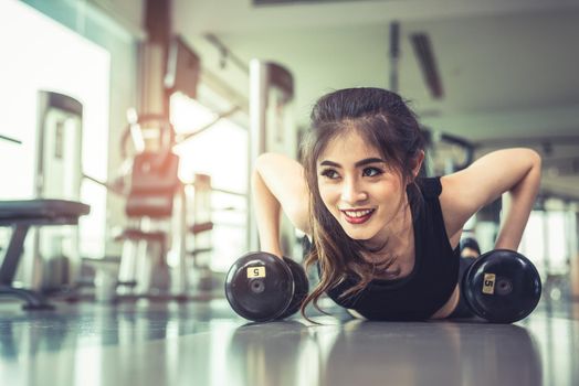 Asian young woman doing push ups with dumbbell on floor in fitness gym and equipment background. Workout and Sport Exercise concept. Healthy and Happiness concept. Beauty and Body build up theme.