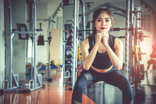 Young Asian woman doing squat workout for fat burning and diet in fitness sports gym with sports equipment in background. Beauty and body build up concept. Sports club and Aerobic theme.