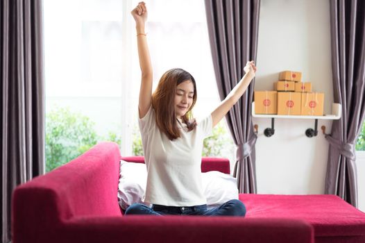 Happy young Asian business woman sitting on red sofa and stretching arm in her house in the morning. Business and Entrepreneur lifestyles concept. Online shopping homepage owner theme.