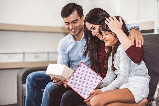 Happy family parents and little girl looking into gift box in Christmas and New year day on sofa in living room. Xmas present for surprised good children in happy home. People and lifestyles concept.